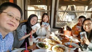 Six people smiling around a sunlit restaurant table sharing plates and drinks.